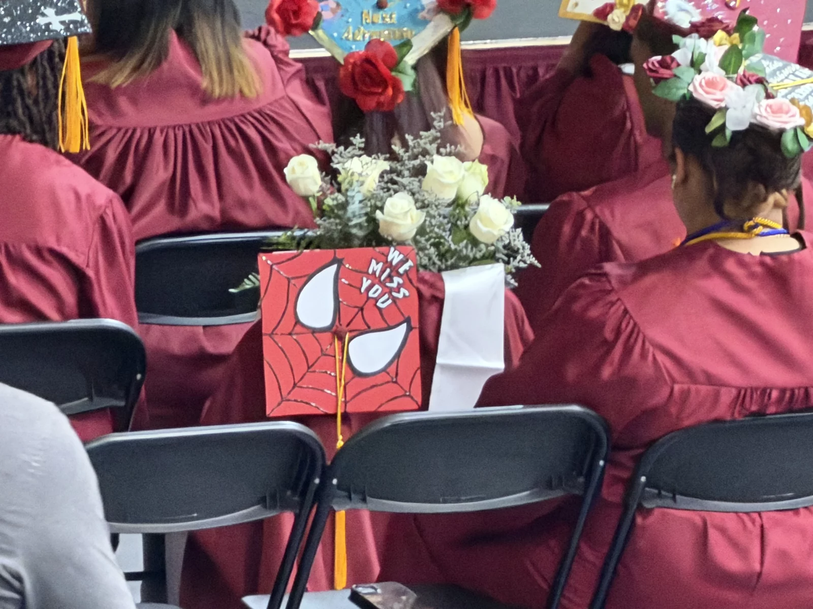 An empty chair with a Spider-Man graduation cap, white roses, and 'We Miss You' message.