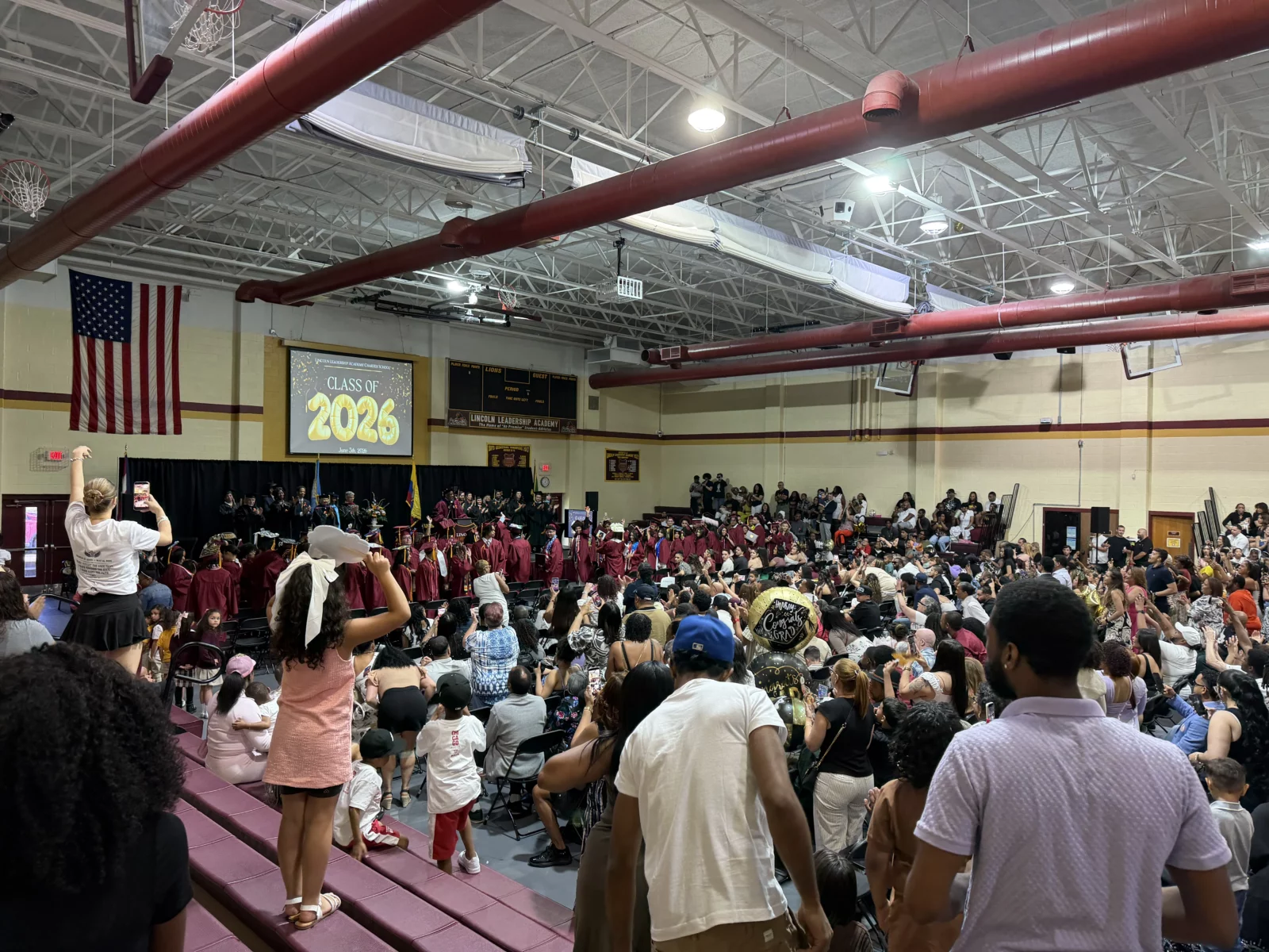 Audience members watch graduates on stage in a gymnasium with a 'Class of 2026' screen.