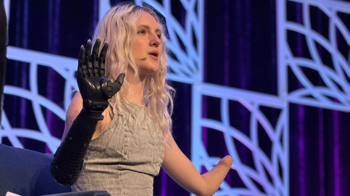 Blonde woman on stage speaks into a microphone, raising her left prosthetic arm.
