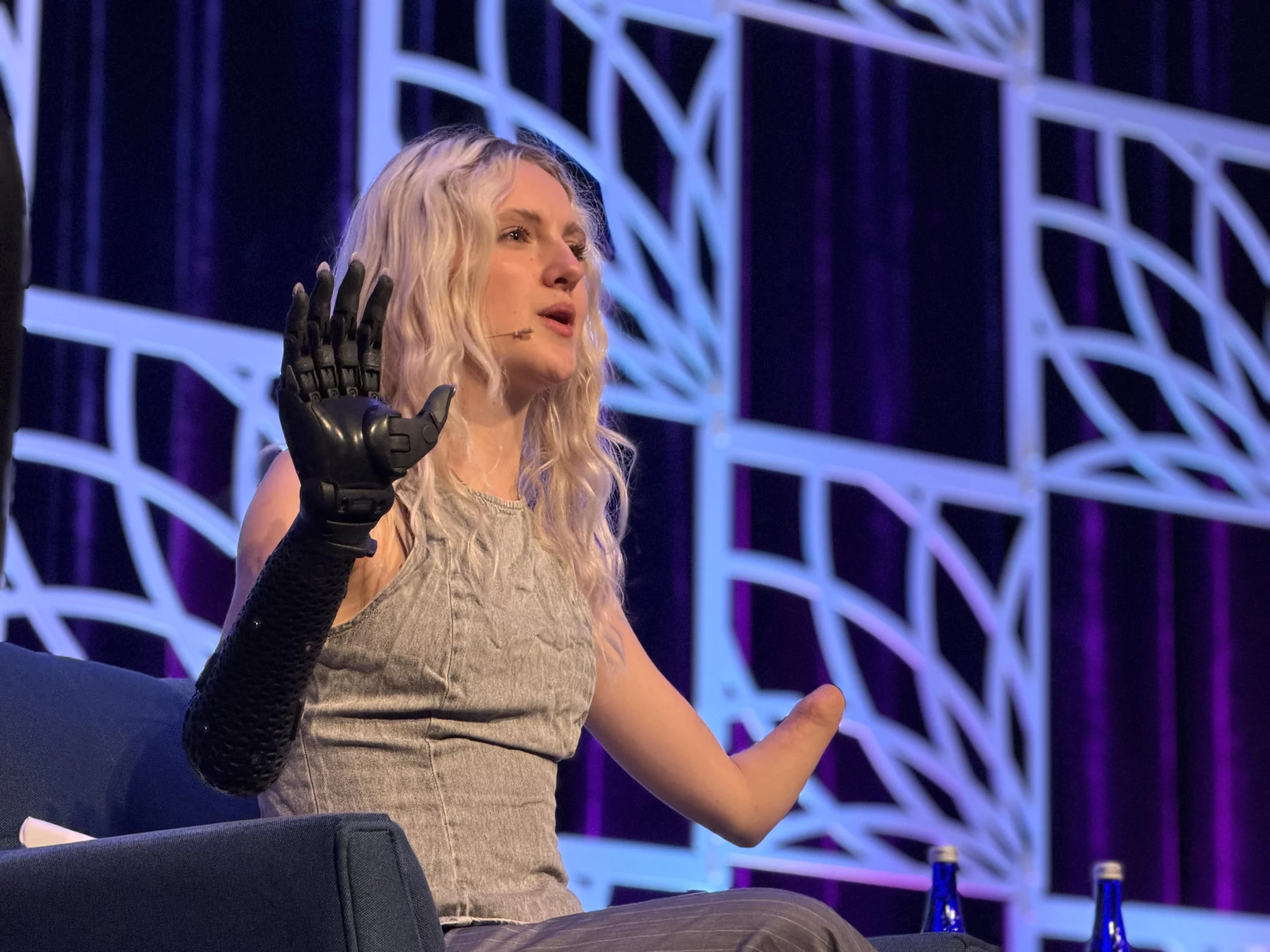 Blonde woman on stage speaks into a microphone, raising her left prosthetic arm.