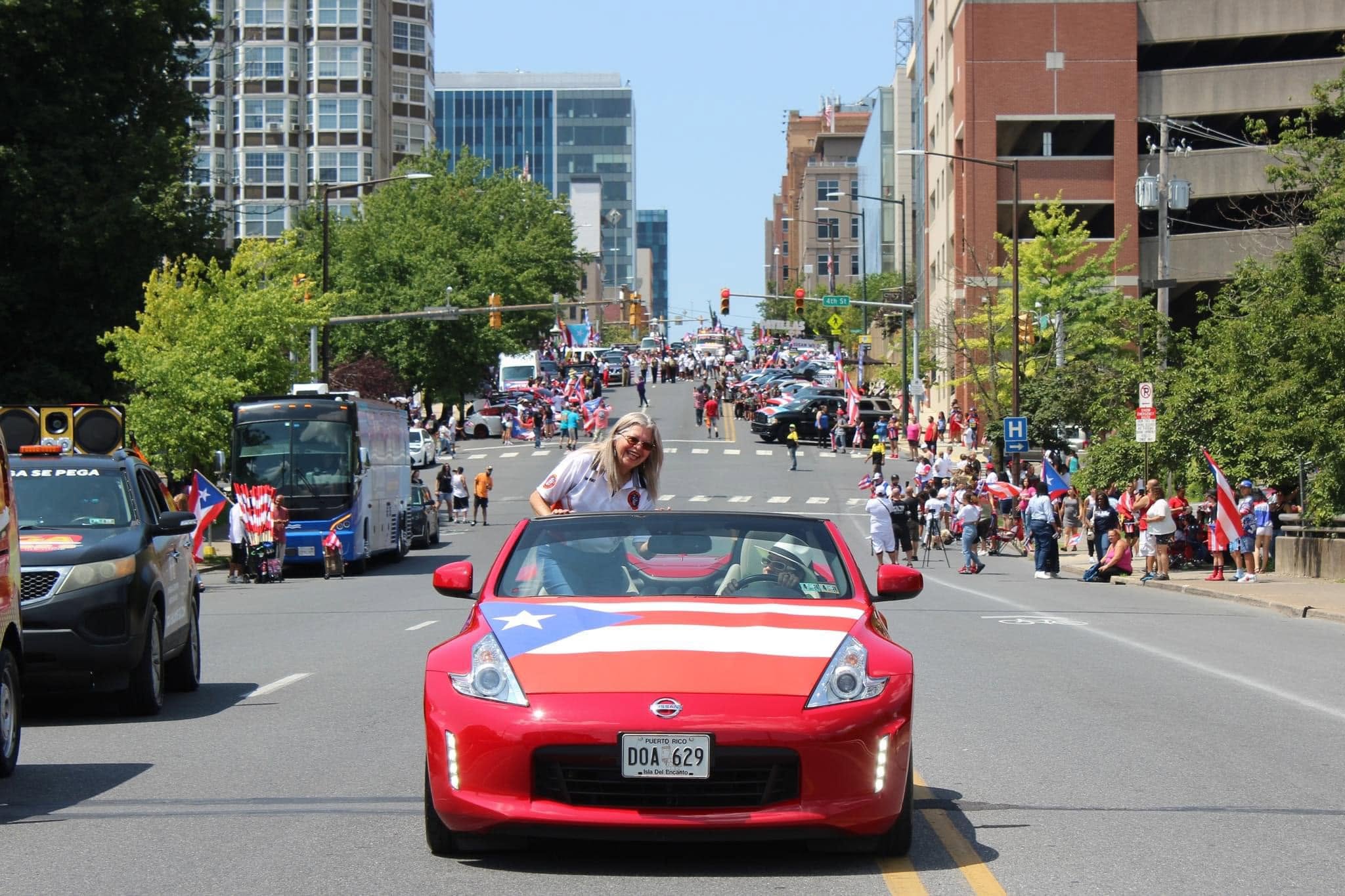 A woman waves from a red convertible decorated with a Puerto Rican flag on a street.