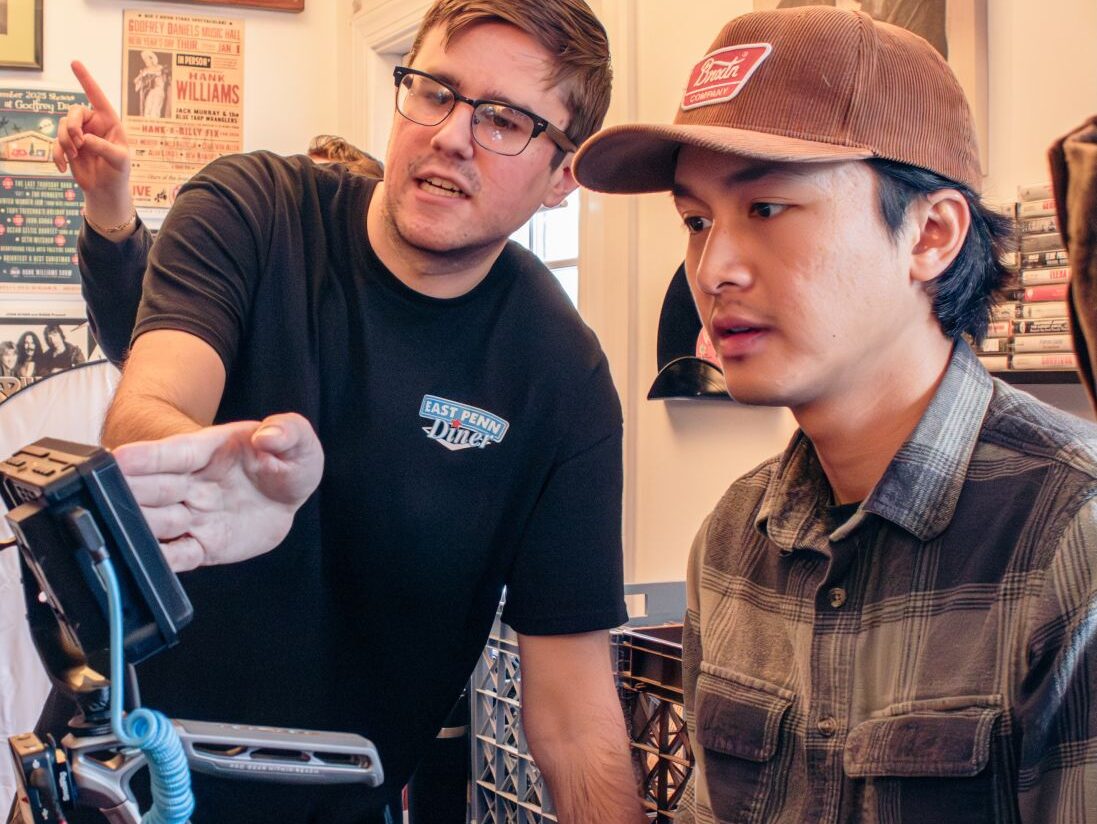 A man wearing glasses points at a camera monitor while standing next to a man wearing a baseball cap.