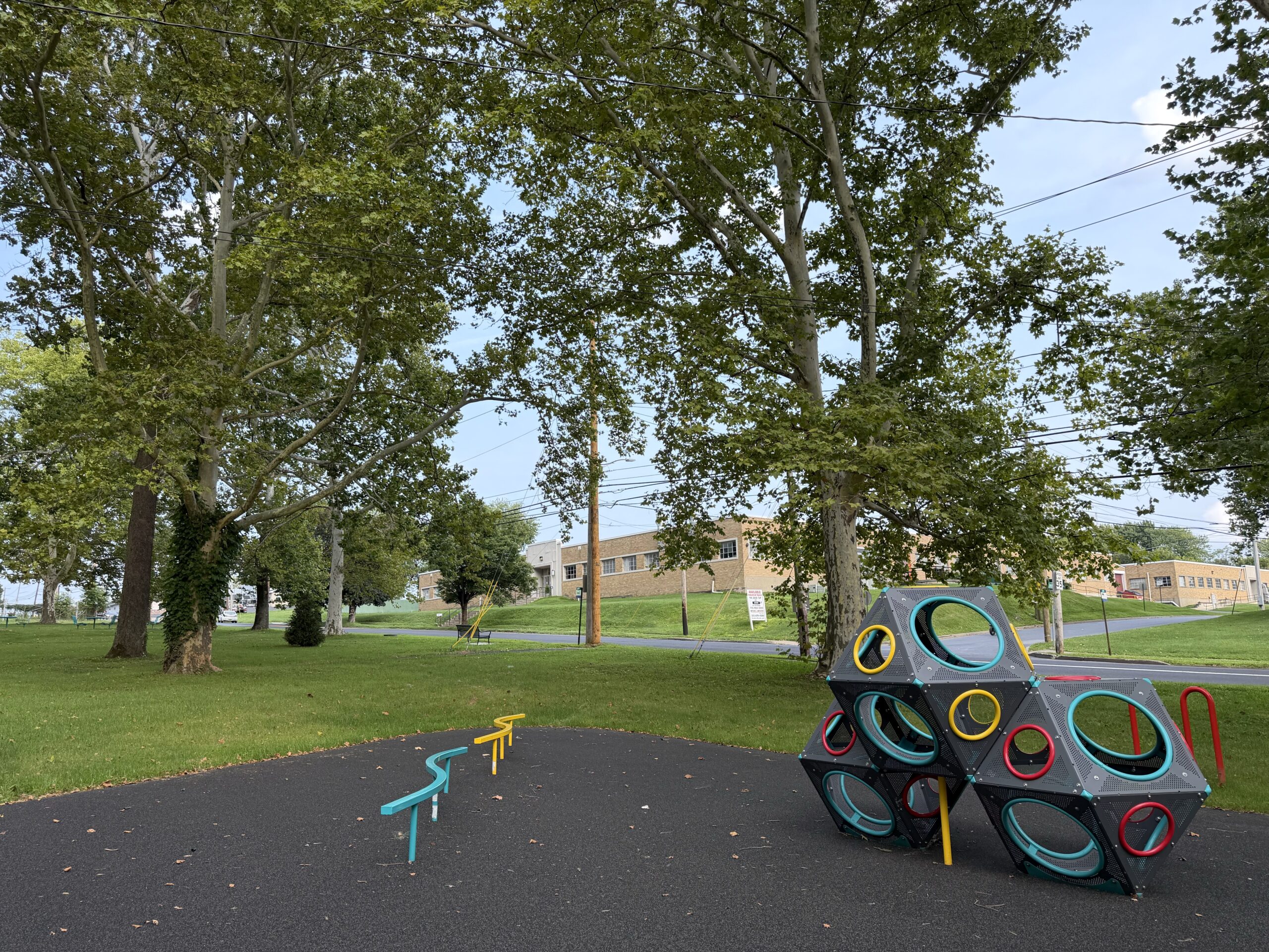 A modern climbing structure with colorful rings sits on a playground under large shade trees.
