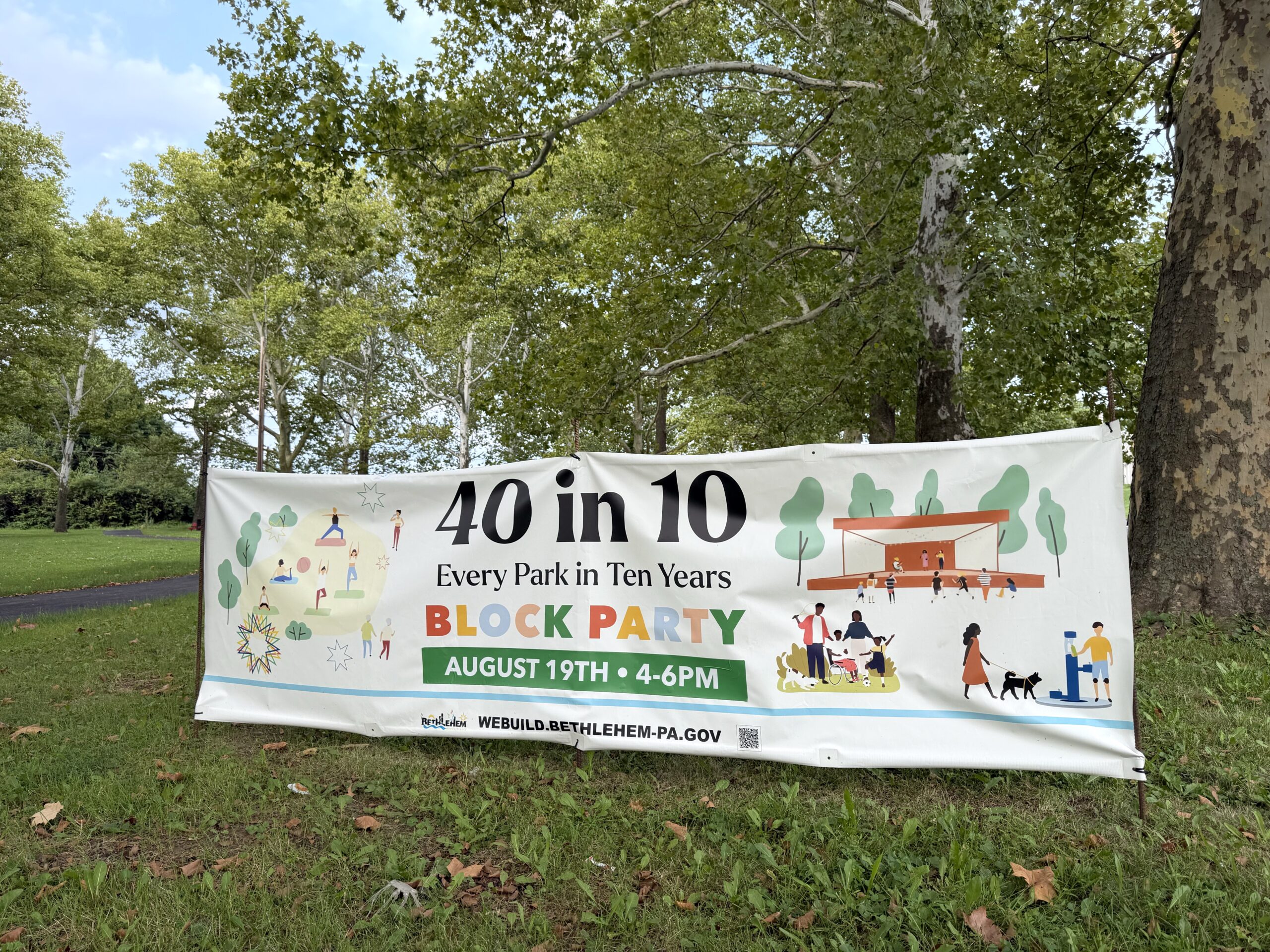 A large white promotional banner stands on a grassy lawn surrounded by trees in a park.