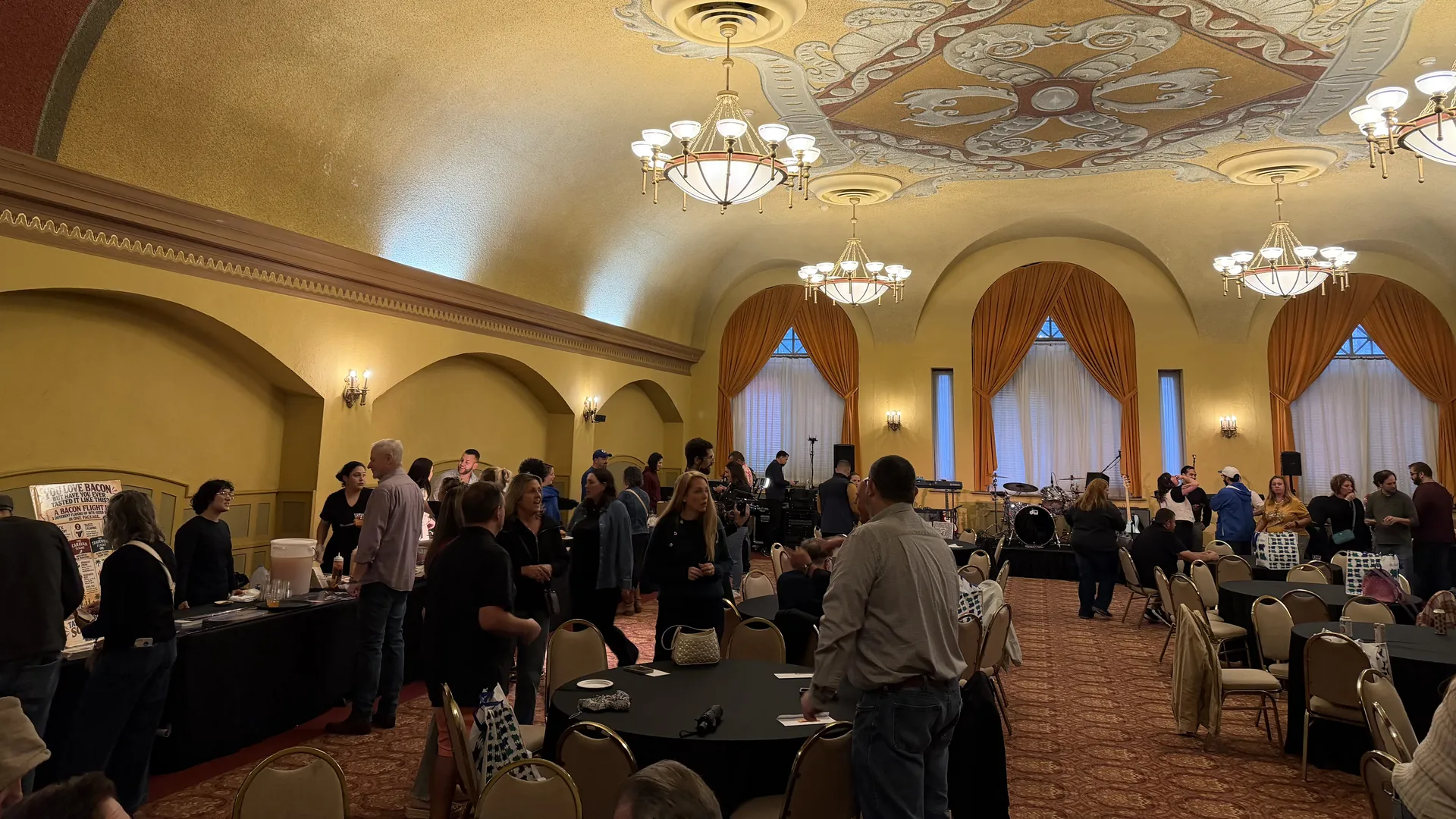 A crowd mingles between vendor tables and round tables in an ornate ballroom with chandeliers, arched windows and orange drapes, with a band setup at the far end.