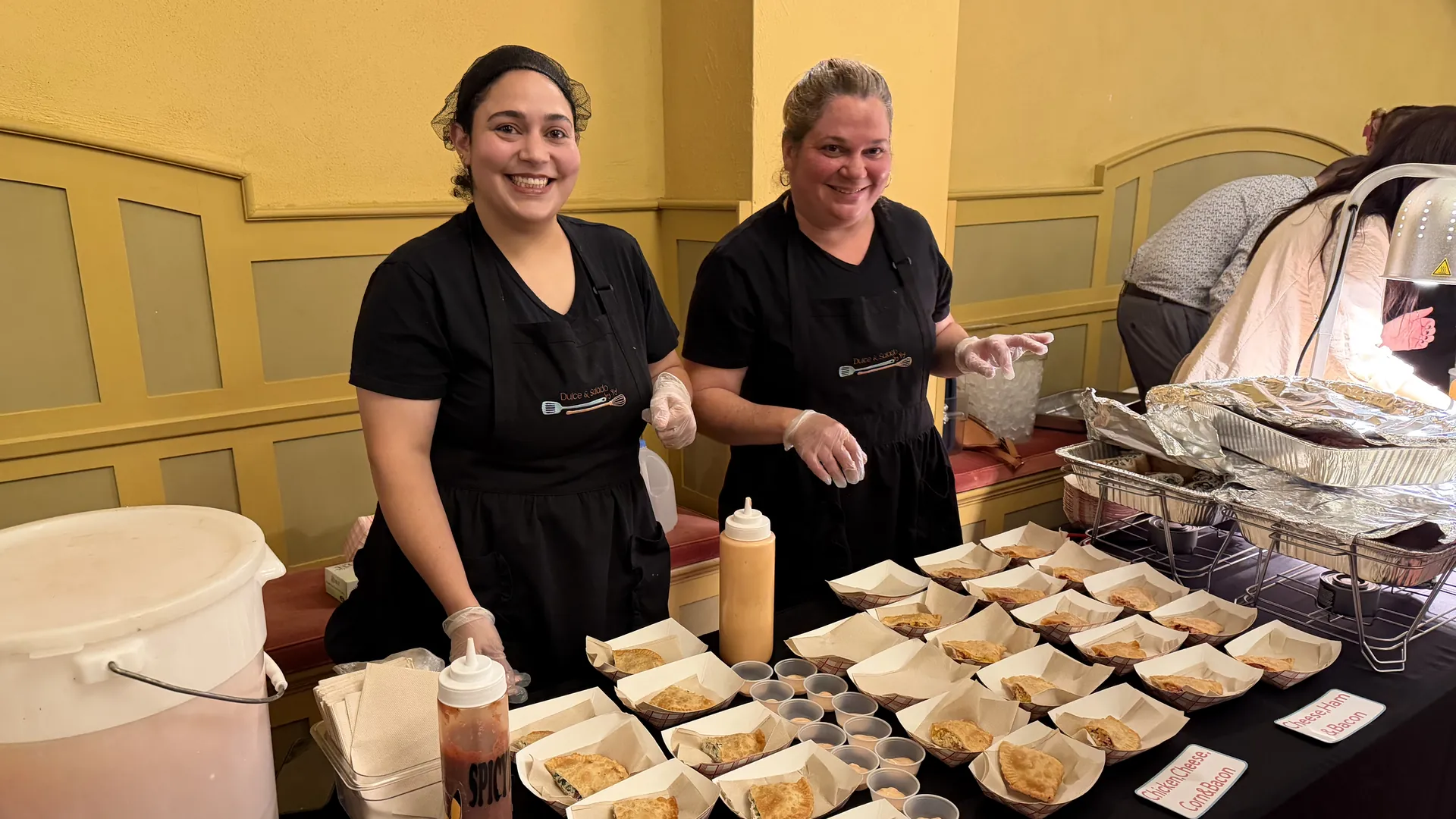 Two smiling women in black aprons stand behind a table lined with empanadas in paper trays and small cups of sauce.