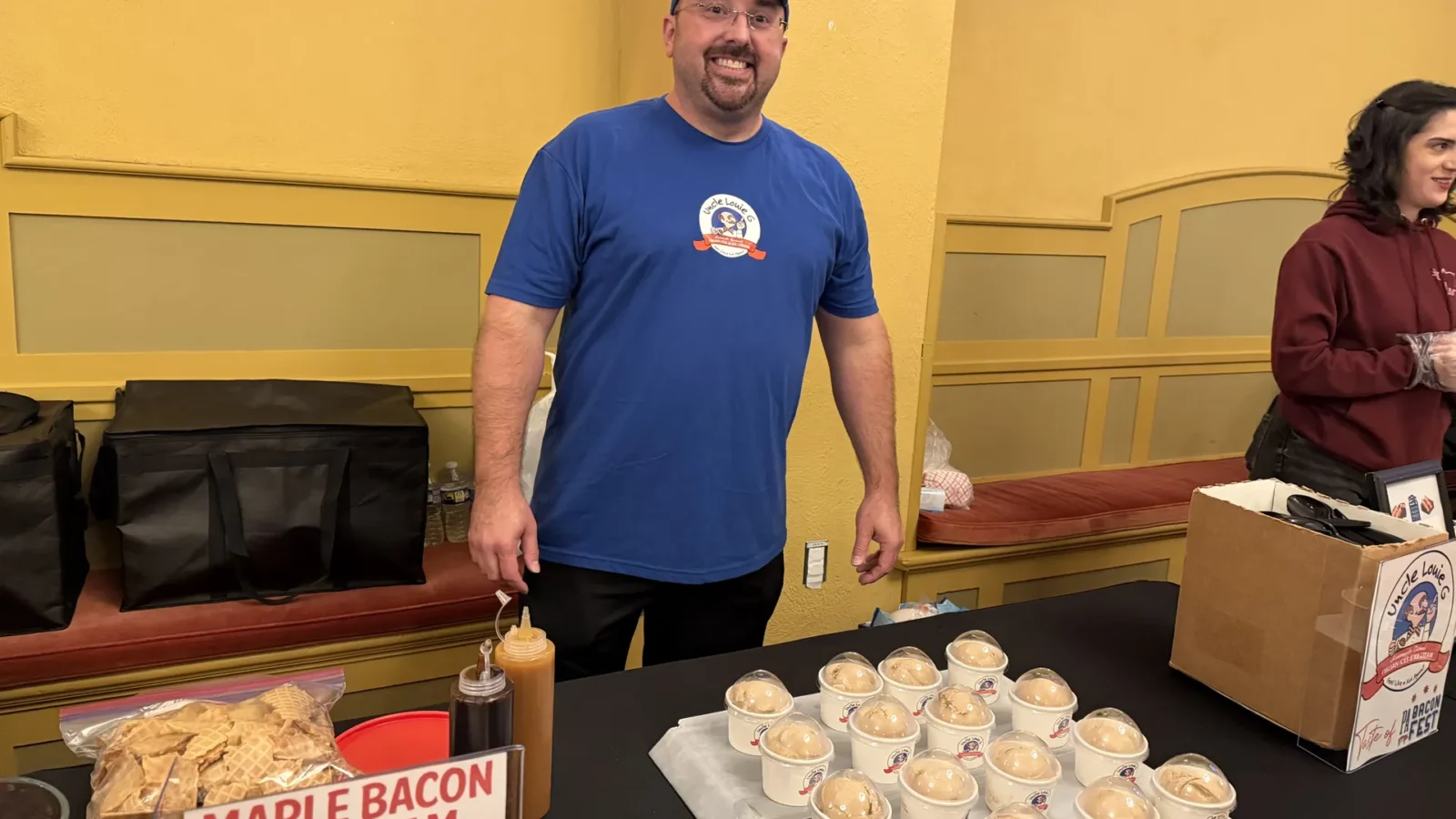 A smiling man in a blue Uncle Louie G T-shirt stands behind a tray of covered ice cream cups beside a Maple Bacon Ice Cream sign.