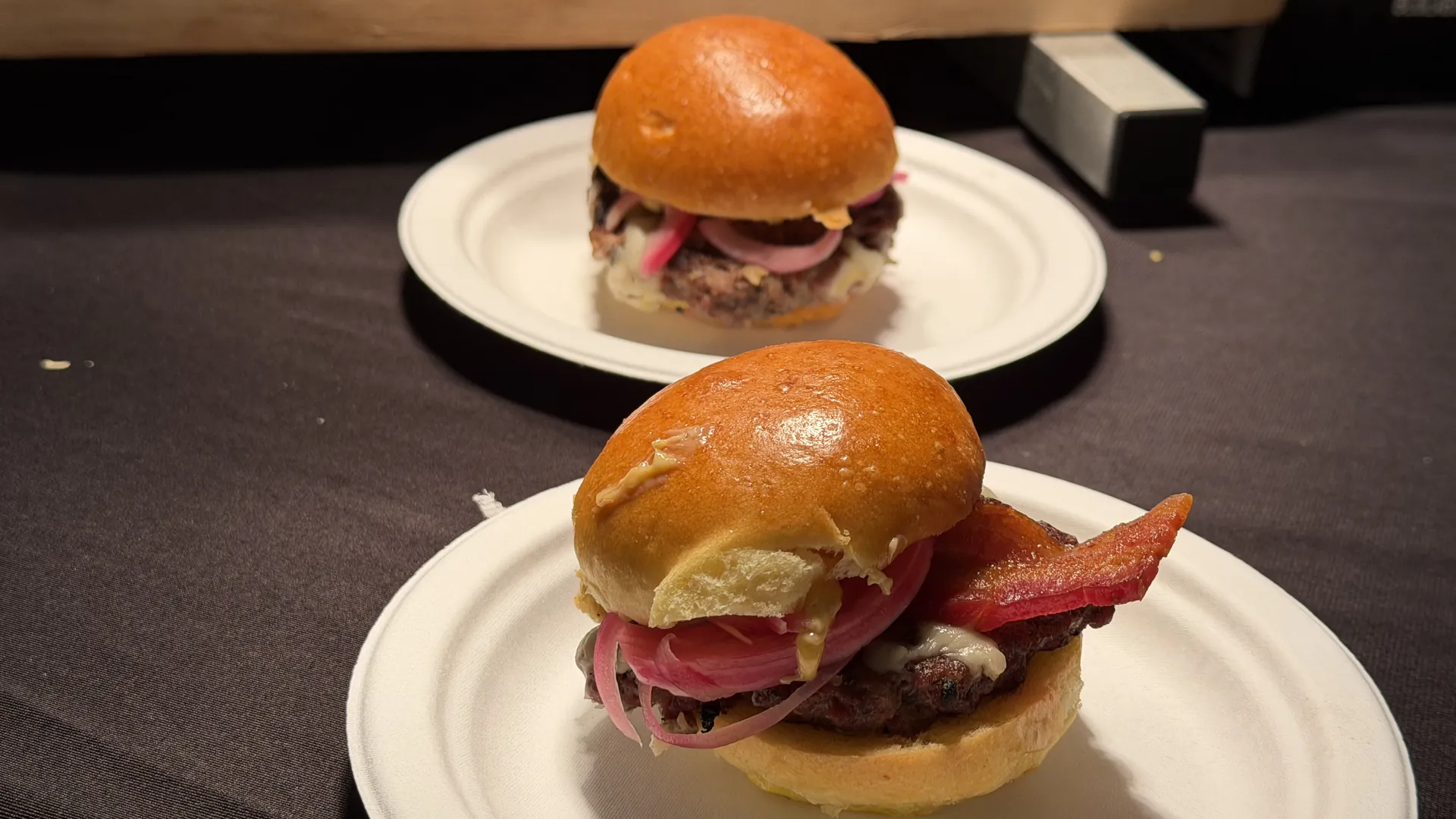 Two small burgers on paper plates, the front one topped with a strip of bacon, pickled red onion and melted cheese on a glossy brioche bun.