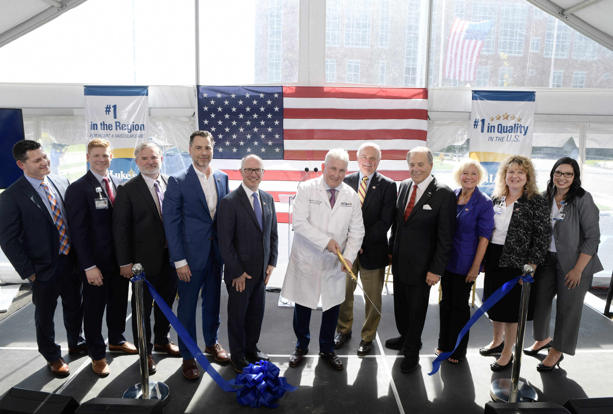 A doctor in a white coat cuts a blue ribbon with oversized gold scissors as 10 people in business attire stand beside him on a stage under a tent, with a large American flag behind them.