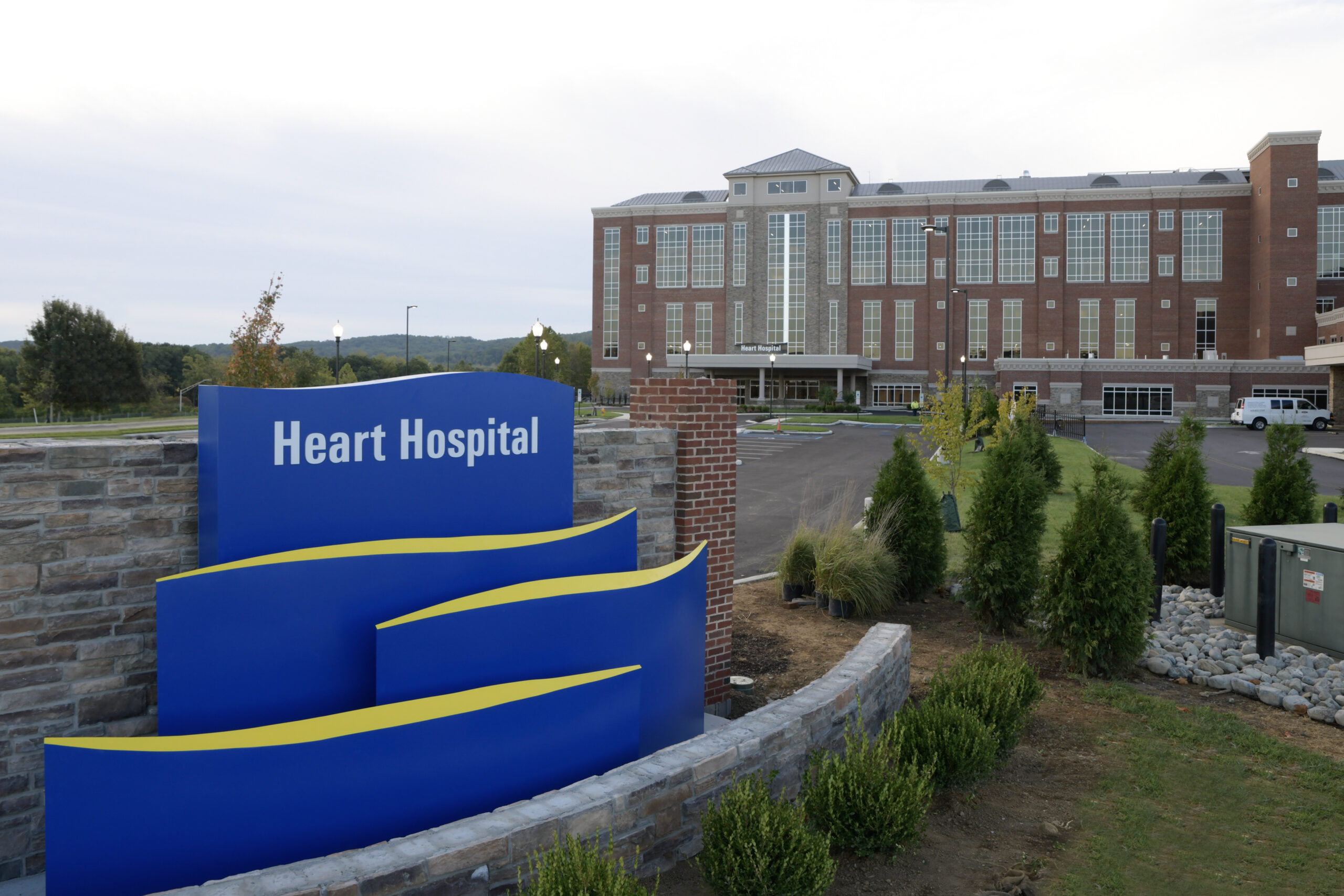 A blue "Heart Hospital" sign on a stone wall stands in front of a large brick hospital building with a covered main entrance, a parking lot, newly planted shrubs and wooded hills in the background.