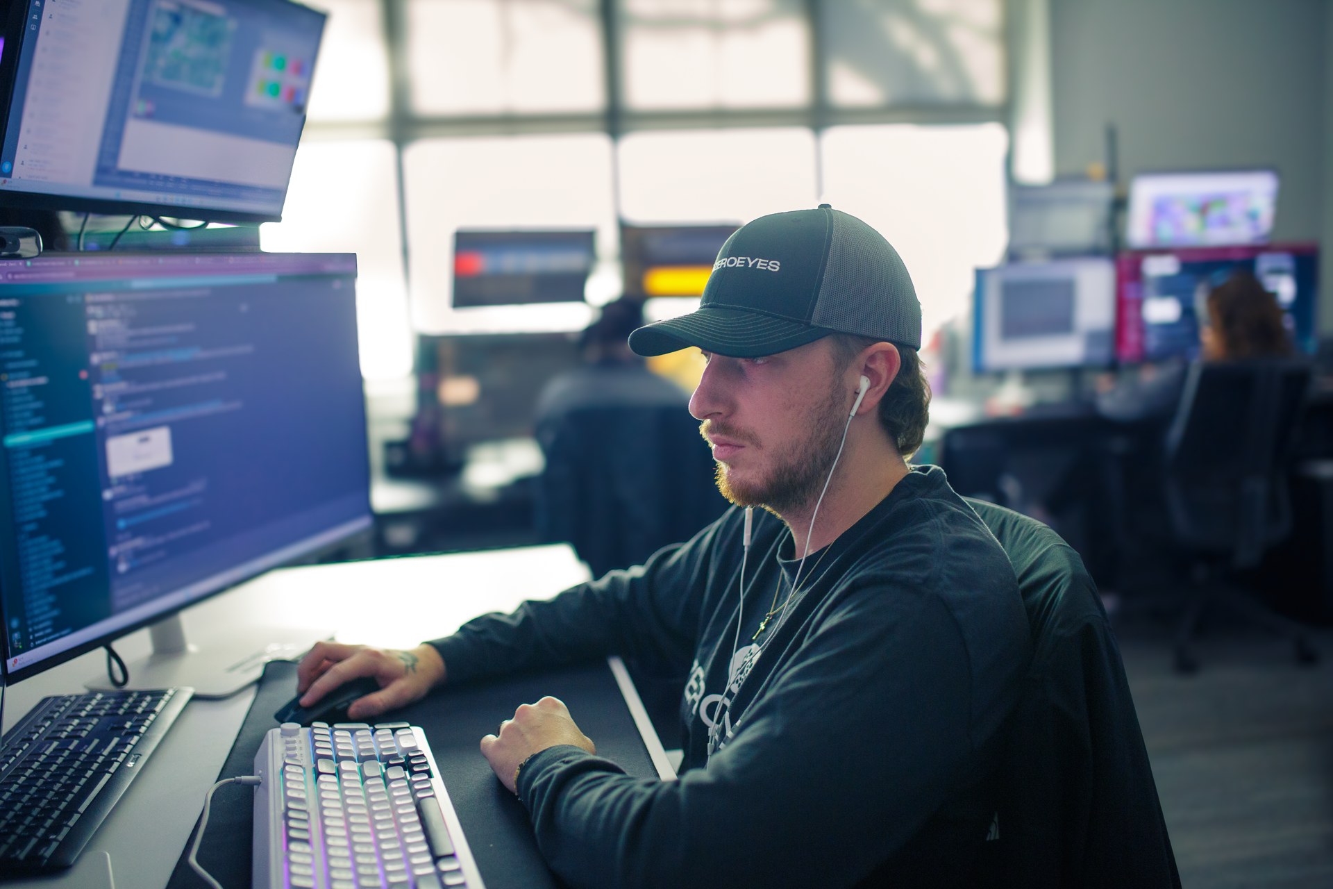 A man in a ZeroEyes cap works at a desk with two computer monitors, with other workers at screens behind him.