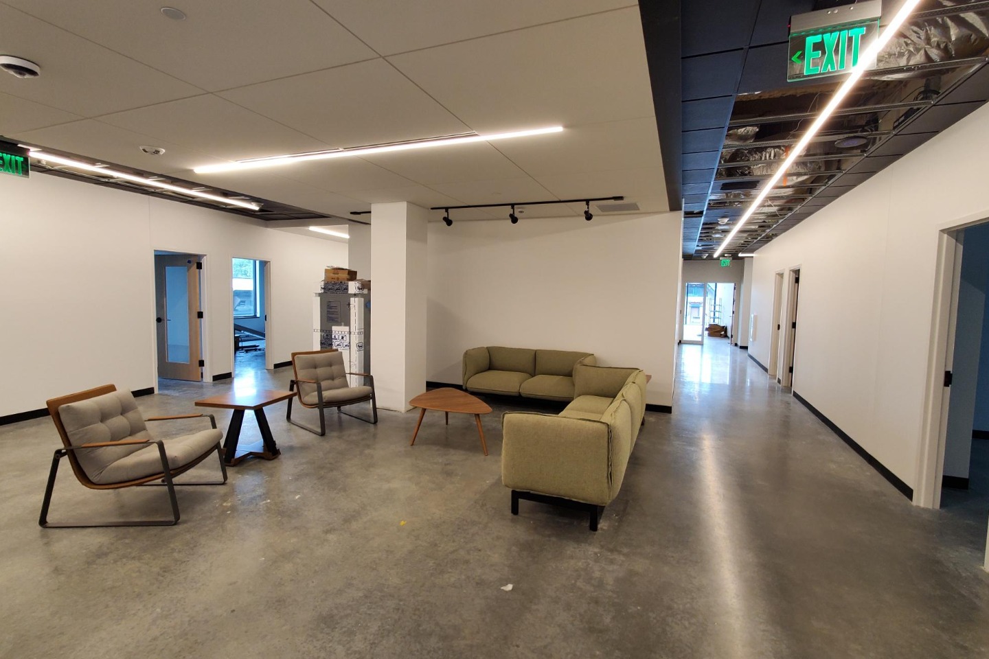 Chairs, a sofa and small tables sit in a lounge area off a hallway with concrete floors and white walls.