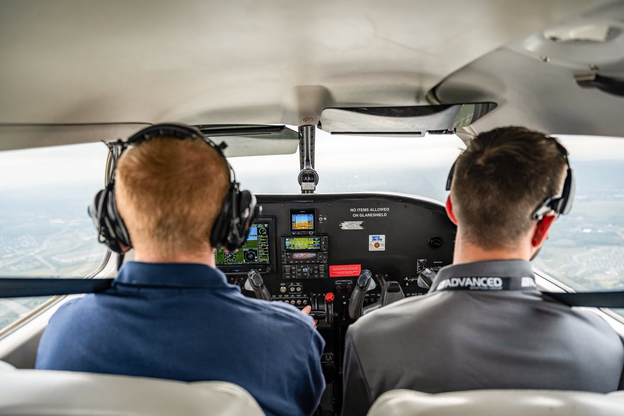 View from the back seat of a small plane in flight, showing two pilots in headsets and an instrument panel with digital screens.
