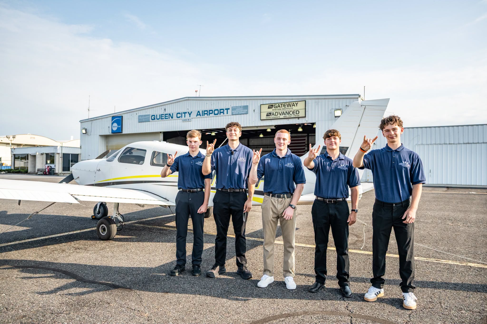 Five students in navy polo shirts stand on the tarmac in front of a small white plane and the Queen City Airport hangar.