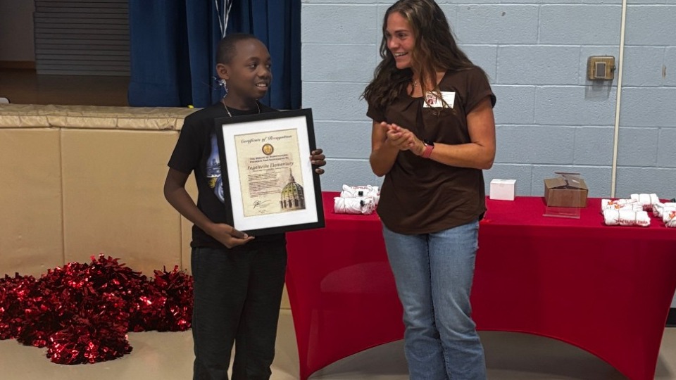 A woman in a brown shirt claps beside a student holding a framed certificate in a school gym decorated with balloons and red pompoms.