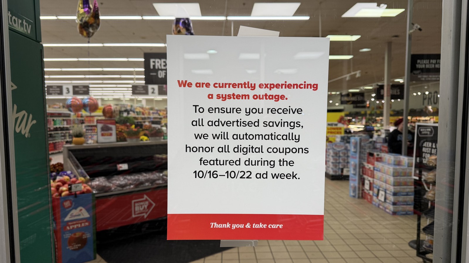 A sign taped to a glass door reads "We are currently experiencing a system outage" above a note on digital coupons, with grocery displays visible inside the store.