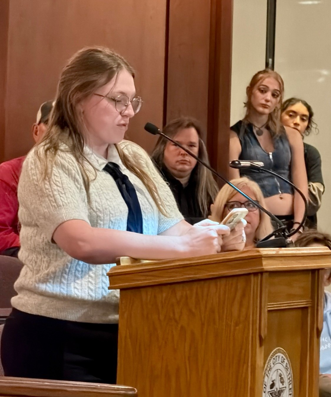A speaker in a cable-knit sweater and tie reads from a phone at a wooden lectern bearing the City of Bethlehem seal as audience members look on.