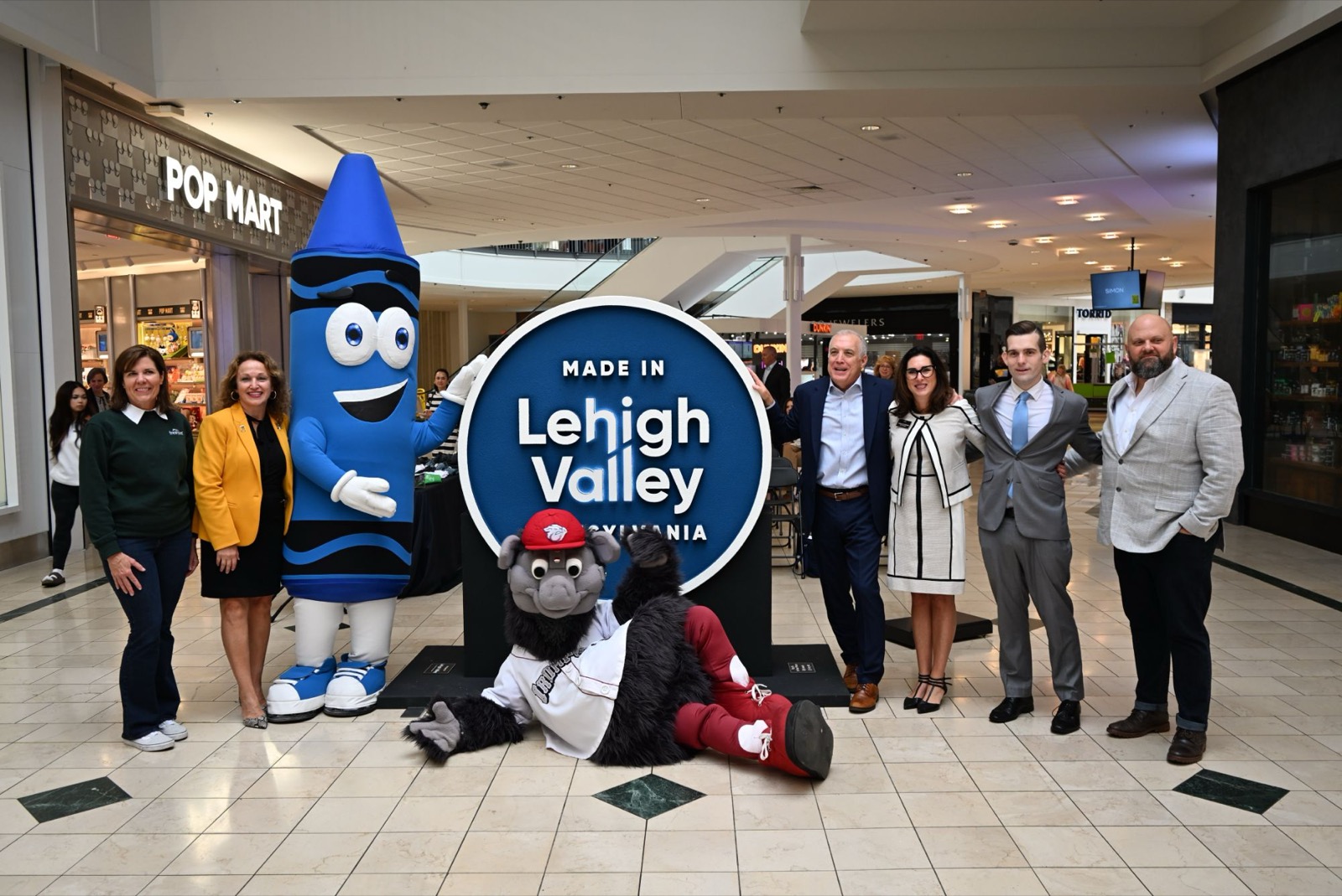 Six adults stand on either side of a large round blue "Made in Lehigh Valley" sign in a mall corridor, with a crayon mascot standing beside it and a furry mascot lying on the floor in front.