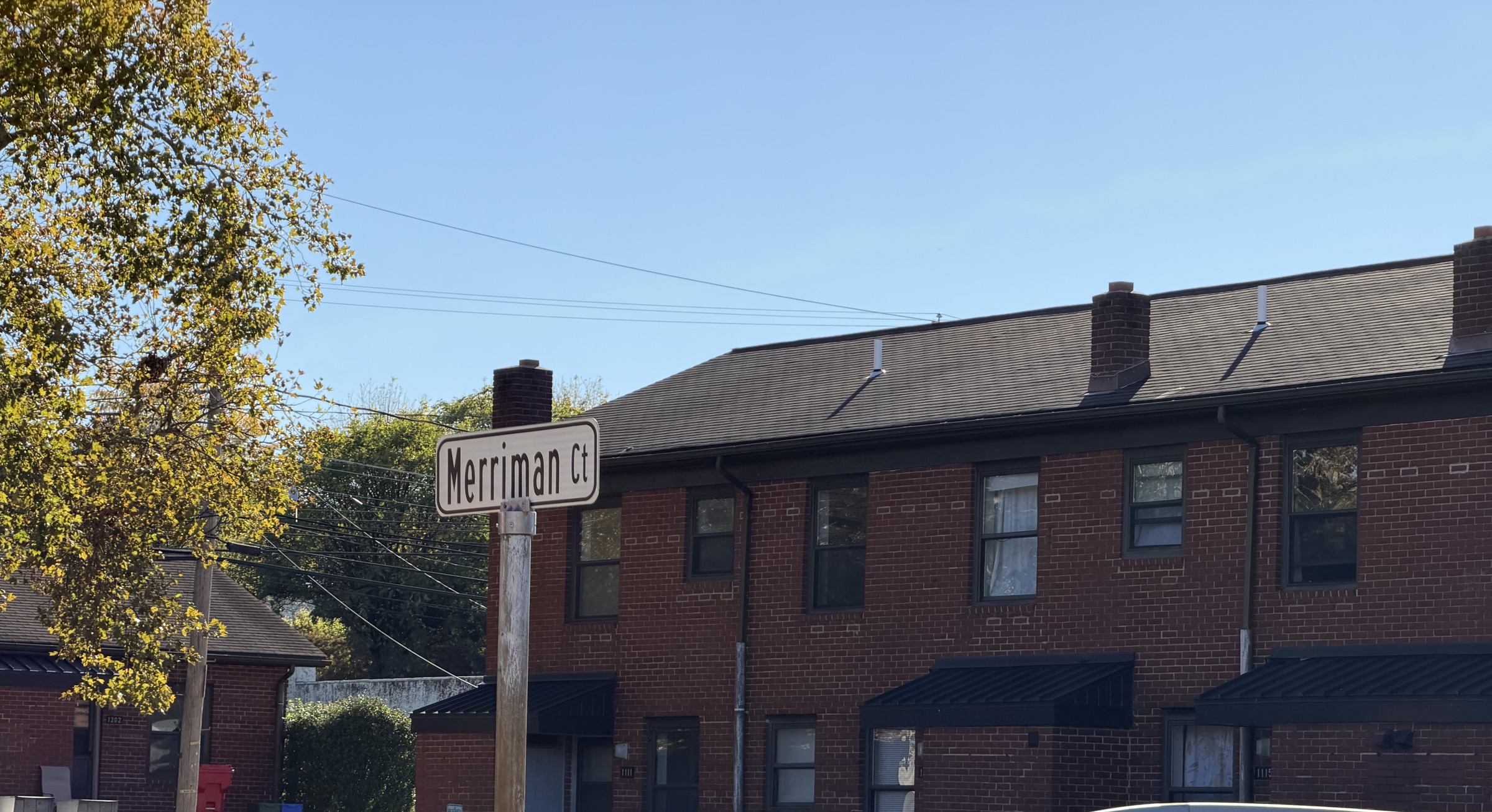 A Merriman Ct street sign stands in front of a two-story red-brick row house, with cars parked in a lot below it.