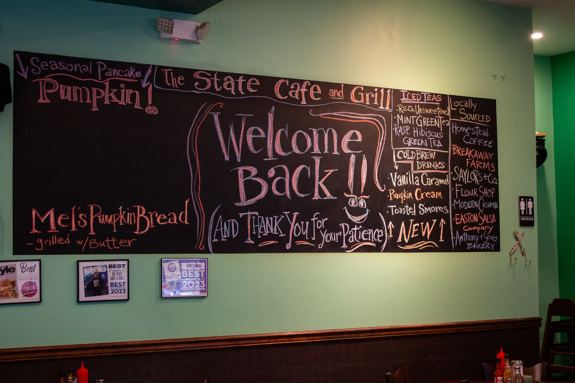 A chalkboard menu wall reading "Welcome Back!! (And Thank You for Your Patience)" with a pumpkin pancake special and seasonal drinks listed.