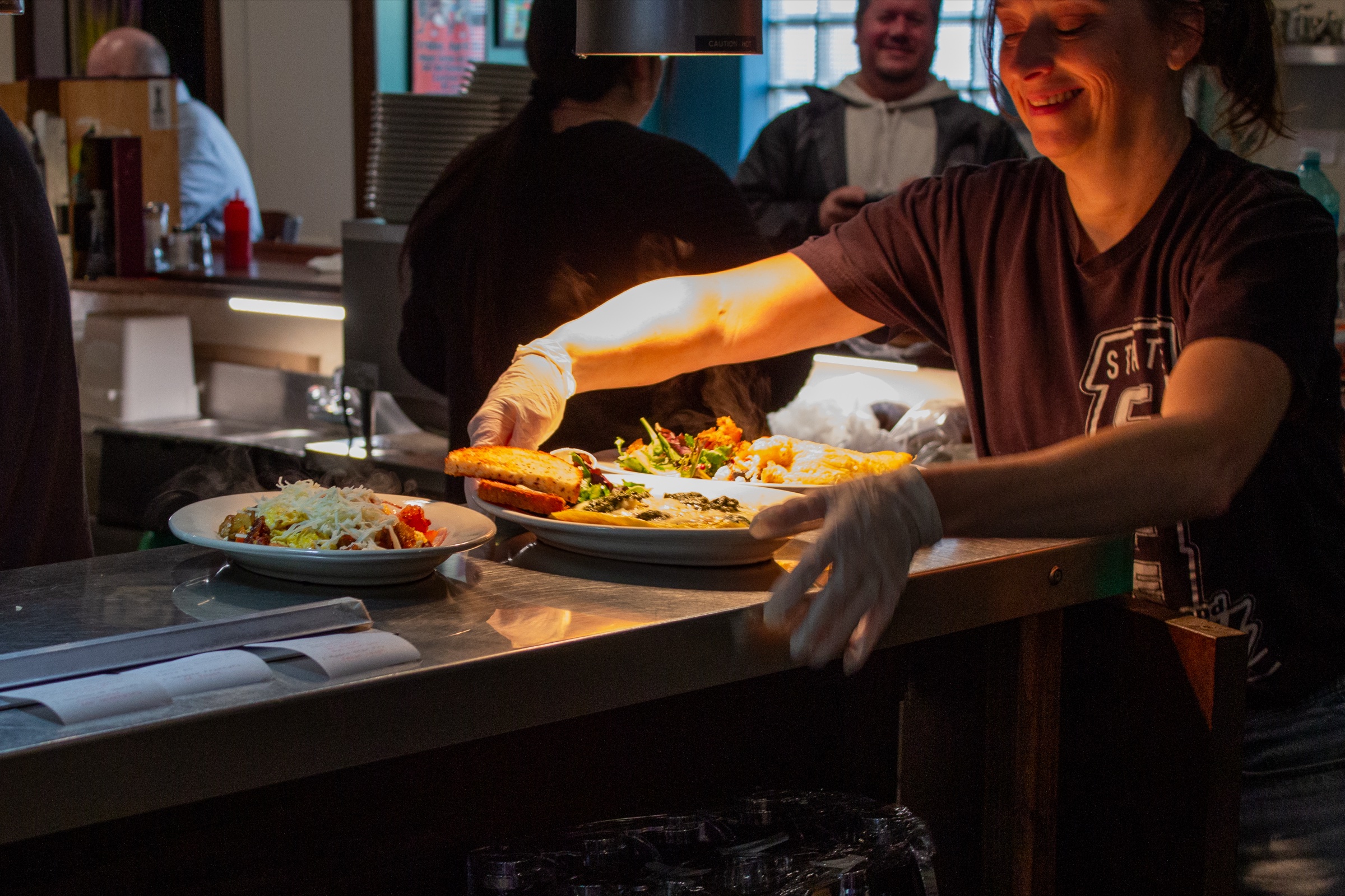 A smiling worker in gloves sets plates of food on the pass in a restaurant kitchen.