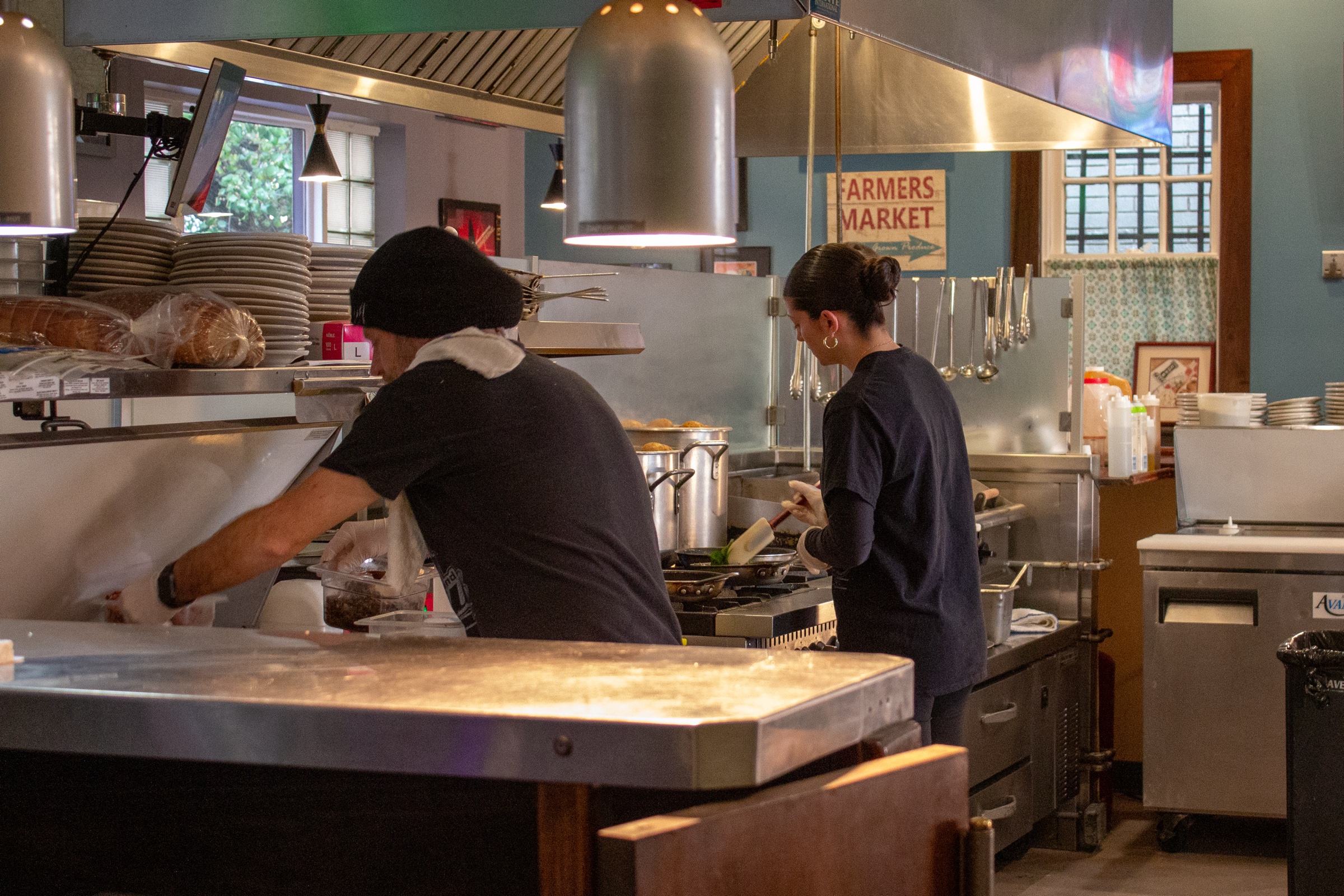 Two cooks work at the grill and prep line in a restaurant kitchen under hanging heat lamps.