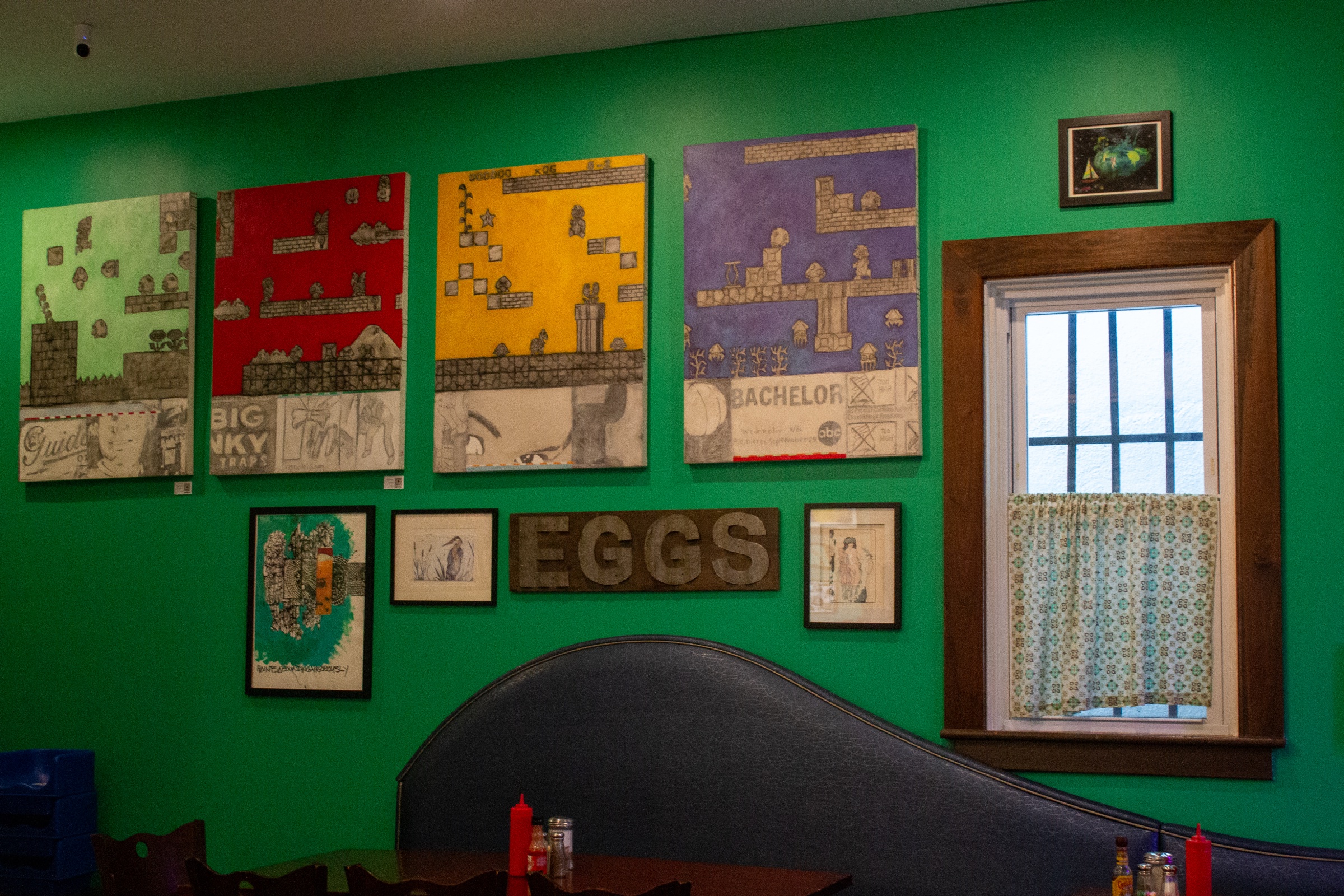 Video-game-style paintings and framed artwork hang on a bright green wall above a dark booth, beside a small window.