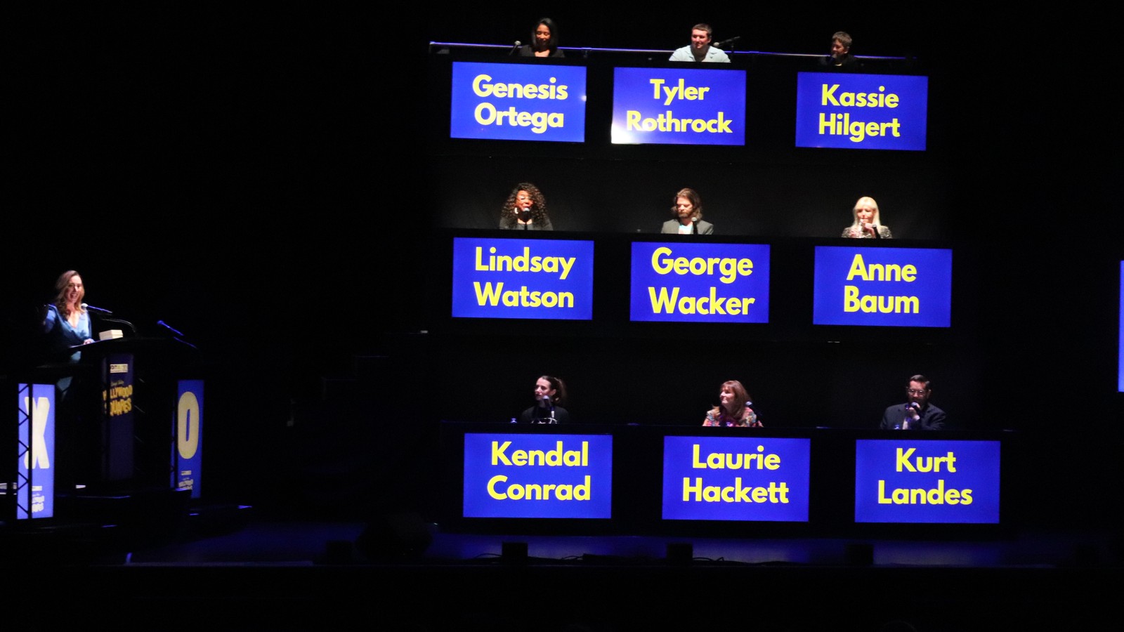 Nine people sit behind blue name placards stacked in a 3-by-3 grid on a dark stage, with a podium at left.